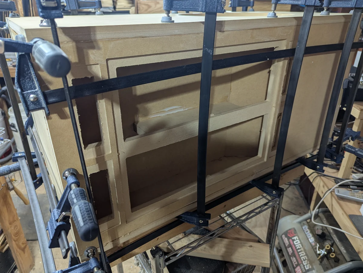 Rear panel of Wanstreet tower speaker cabinet showing two recessed amplifier mounting pockets — upper pocket for 1000 watt mid and high amplifier and lower pocket for 2500 watt subwoofer amplifier — being clamped during assembly