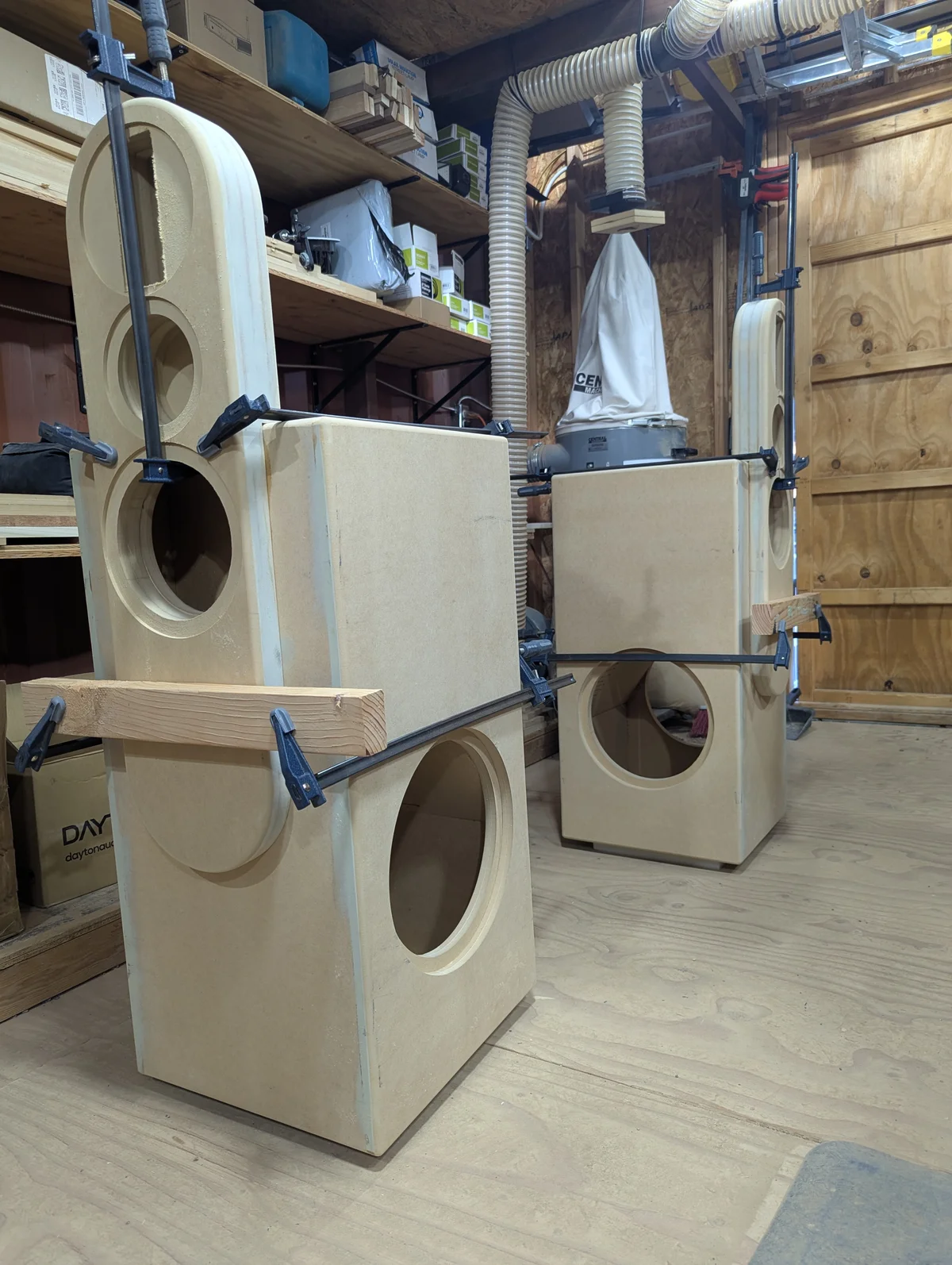 Both Wanstreet tower speaker cabinets assembled and standing in workshop showing pill-shaped upper sections with driver cutouts above large separate subwoofer base enclosures with 12-inch driver openings on front and side faces