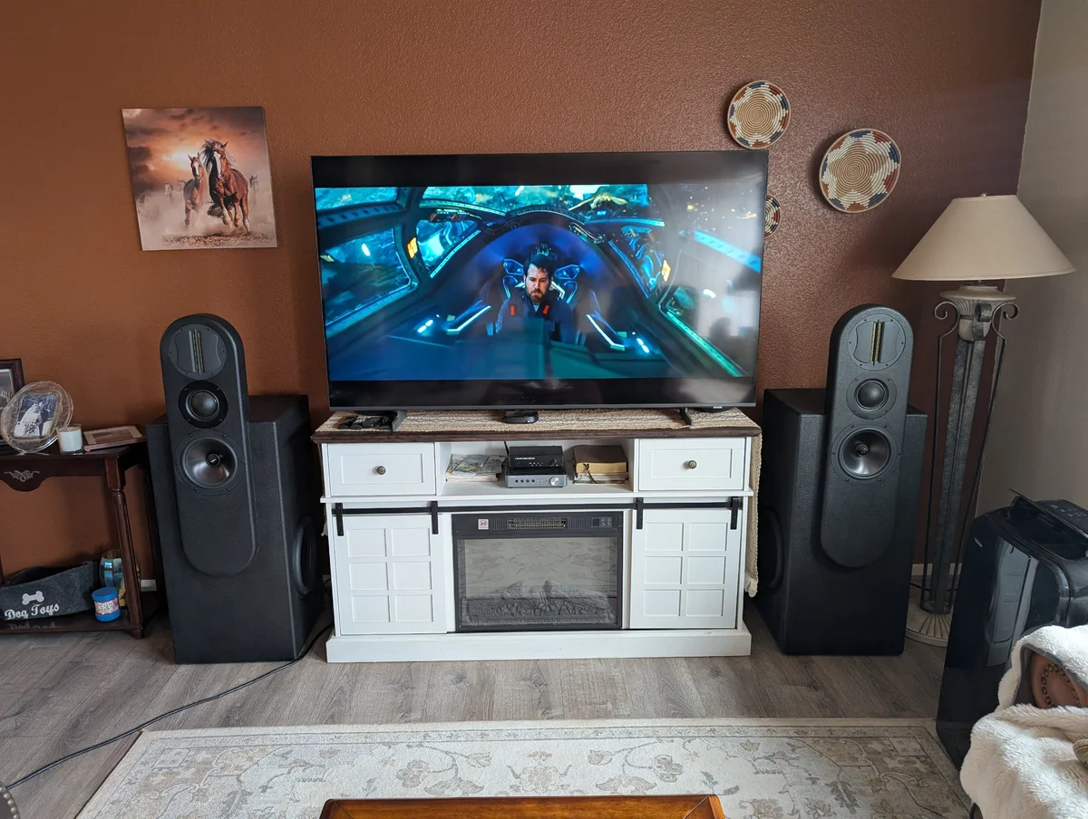 Completed Wanderstreet Scottsdale custom WubWub Audio tower speakers installed flanking a large TV in a living room, showing ribbon tweeter, dome mid, and 8-inch woofer on pill-shaped upper sections with powered 12-inch subwoofer base enclosures, system playing a movie